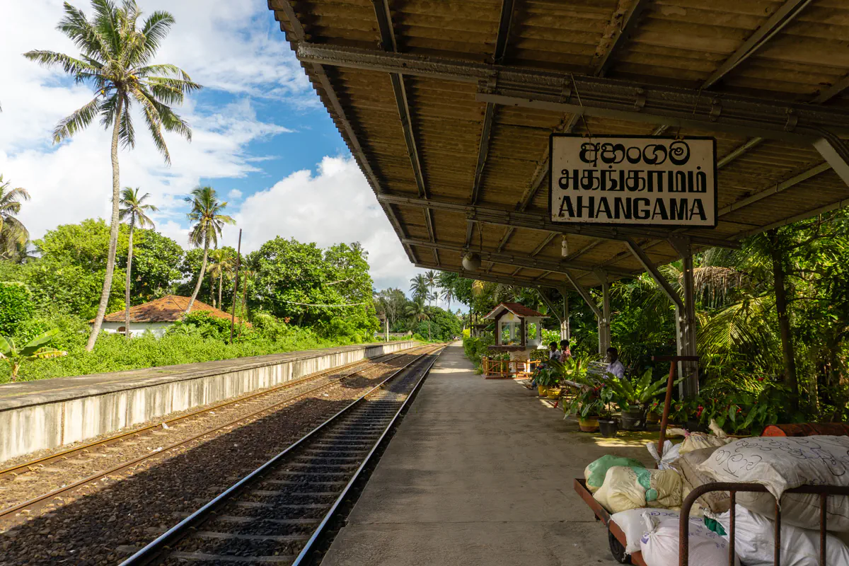 A quiet train platform at Ahangama station with a trilingual sign, surrounded by palm trees and dense greenery under a partly cloudy sky.