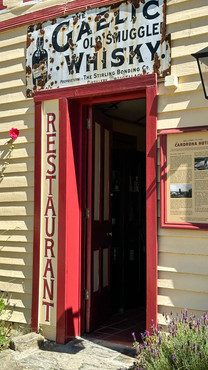 The entrance to the historic Cardrona Hotel features a weathered sign advertising old smuggled whisky. The open red door and vintage exterior highlight the building’s heritage character.