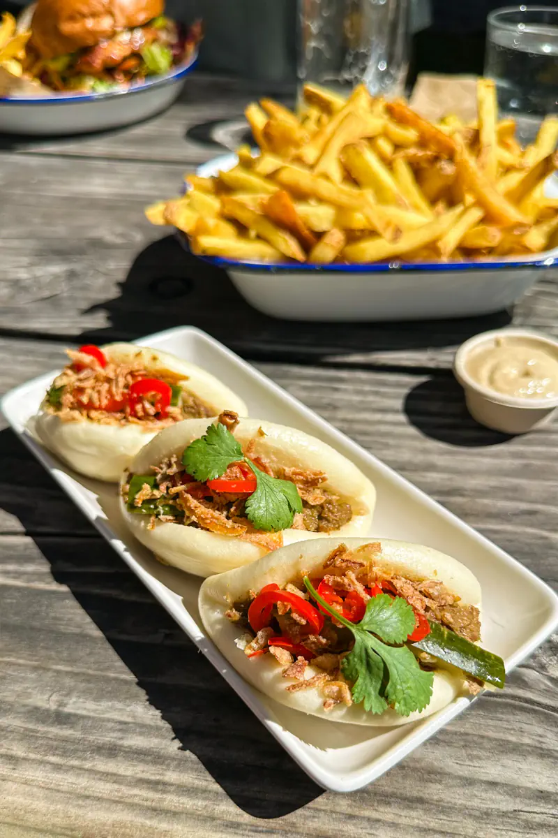 Three steamed bao buns topped with meat, herbs, and sliced chilies sit on a plate beside a bowl of fries. The food is photographed outdoors on a wooden table, suggesting a relaxed pub-style meal.
