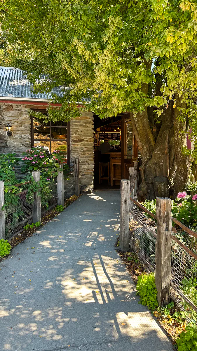 A stone building entrance sits beneath leafy green trees along a narrow path lined with flowers. The shaded walkway creates a cosy, welcoming atmosphere.