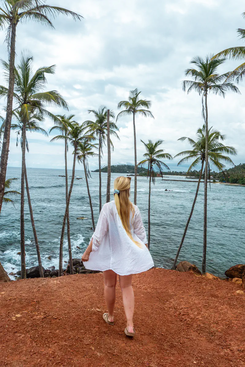 A woman with long blonde hair, dressed in a white cover-up, stands on a red cliff overlooking the ocean, framed by tall, wind-blown palm trees. Waves crash against the rocks below under a cloudy sky, with a small headland visible in the distance.