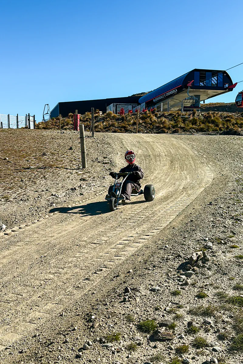A rider wearing a helmet speeds downhill on a mountain cart along a gravel track beneath a gondola station. The alpine terrain and clear blue sky highlight this outdoor adventure experience.