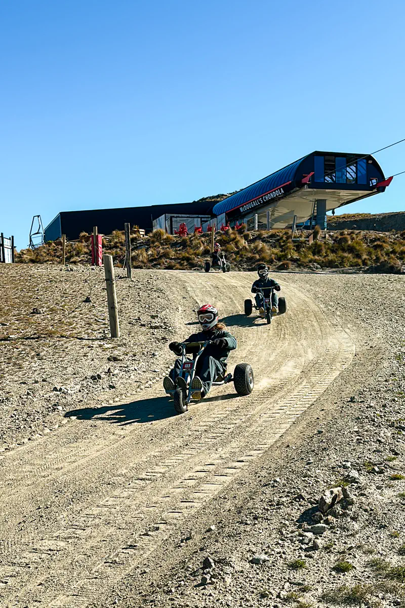 Two riders descend a gravel mountain cart track with a gondola station visible at the top of the hill. The scene captures motion, dust, and the open alpine setting.