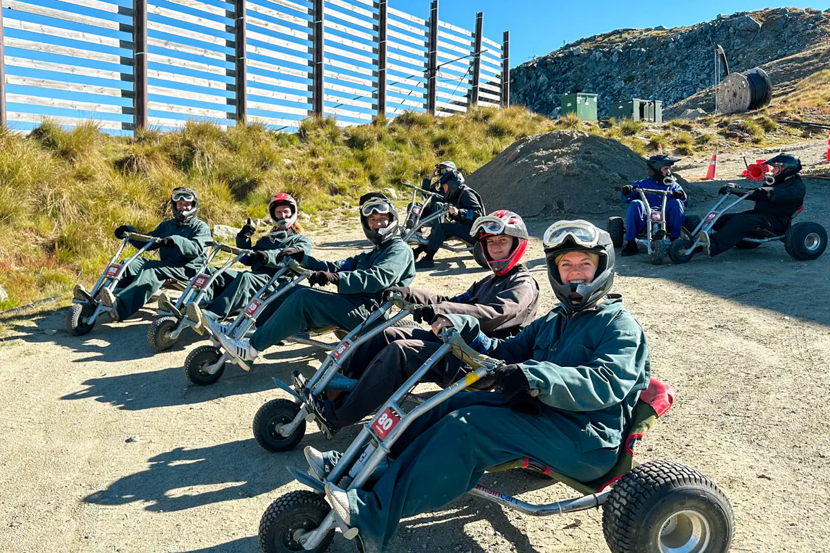 A group of riders sit on mountain carts wearing helmets and protective clothing, lined up at the start of the track. The dry, grassy hillside and fencing frame the staging area.