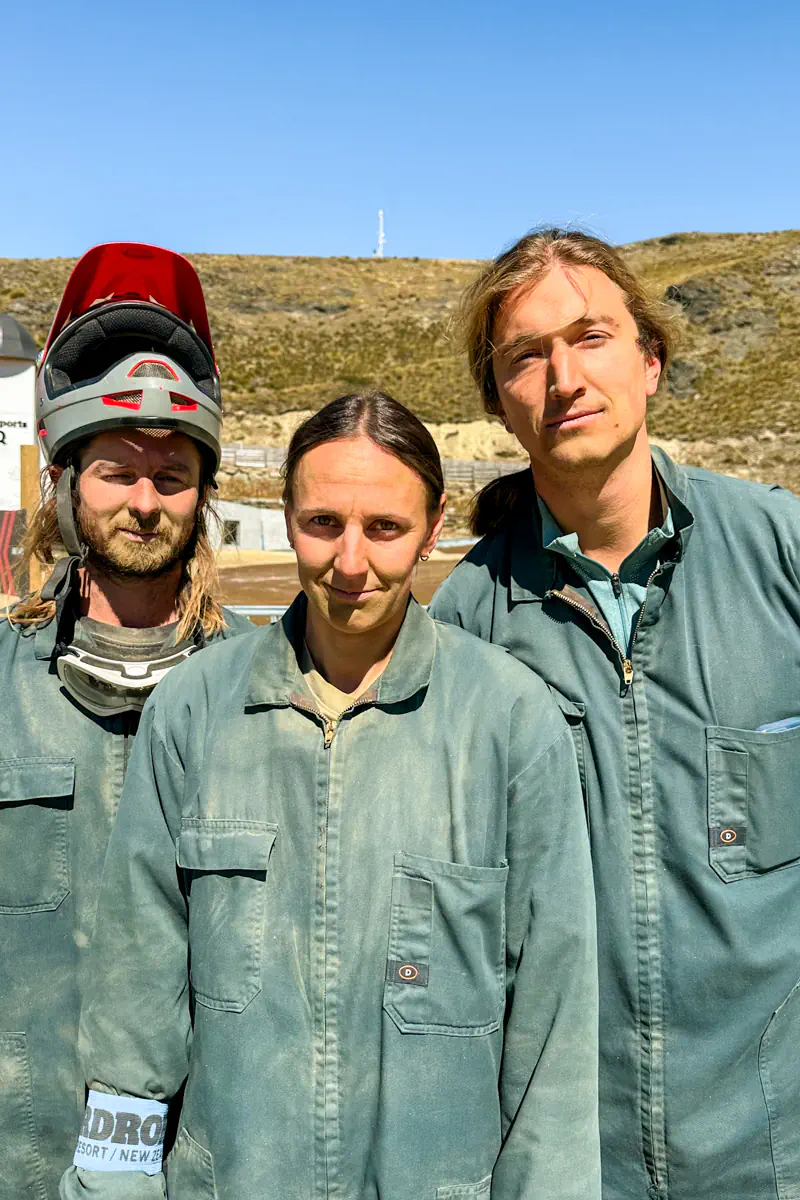Three friends stand together wearing green coveralls and safety gear at an alpine resort. You can see a lot of dust on their faces after mountain carting.