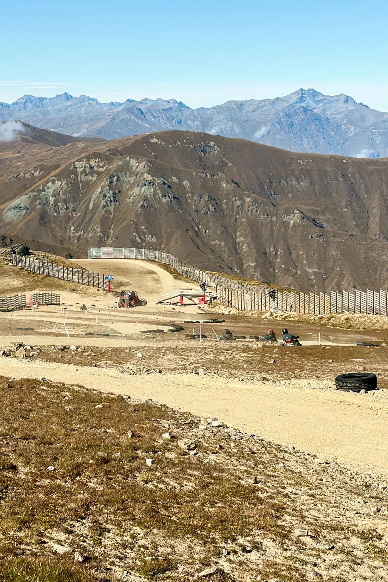 A wide view of a winding mountain cart track cutting across a rugged alpine landscape. Riders descend the trail with dramatic mountain peaks in the distance.