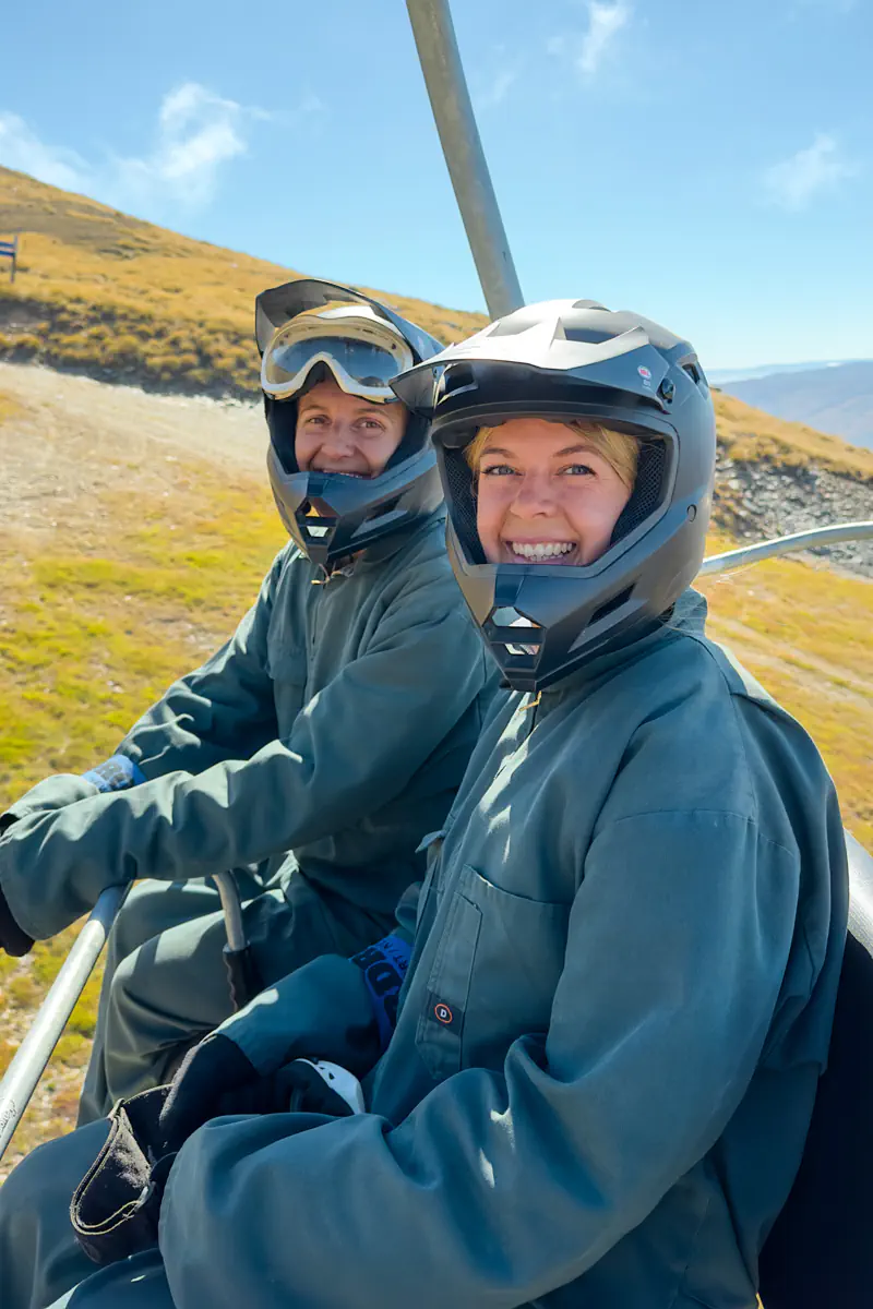 Two people wearing helmets and protective coveralls smile while riding a chairlift up the mountain. The open alpine terrain and sunny conditions suggest they are heading to the start of the carting run.