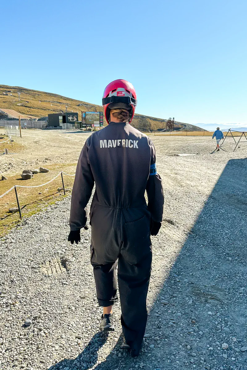A person wearing a helmet and dark coveralls walks across a gravel area toward the mountain carting track. On the back of the coveralls it says "Maverick".