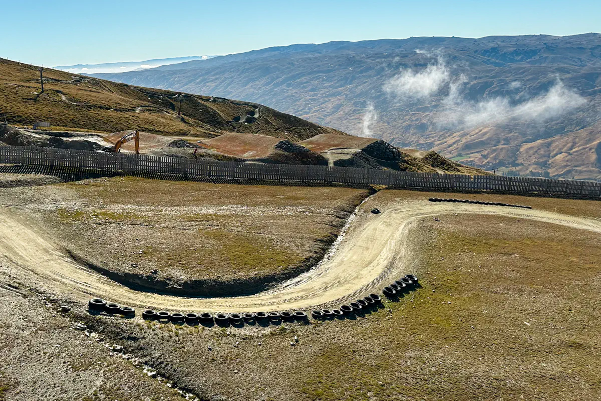 An empty mountain cart track curves downhill with stacked tires lining the corners for safety. Rolling hills and distant valleys surround the course, showcasing the rugged alpine environment.