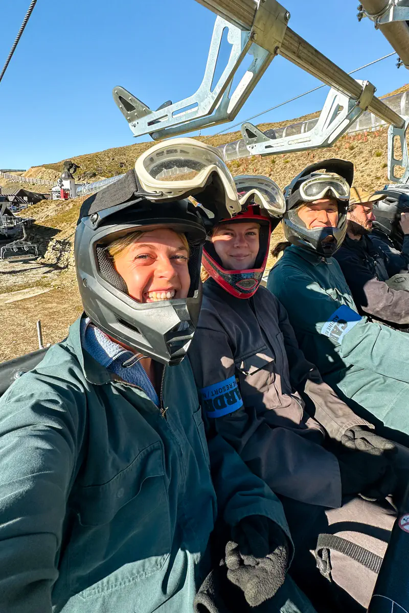 Three people wearing helmets and protective gear sit on a chairlift, smiling as they ride uphill. The lift carries them above the resort terrain toward the start of the mountain carting run.