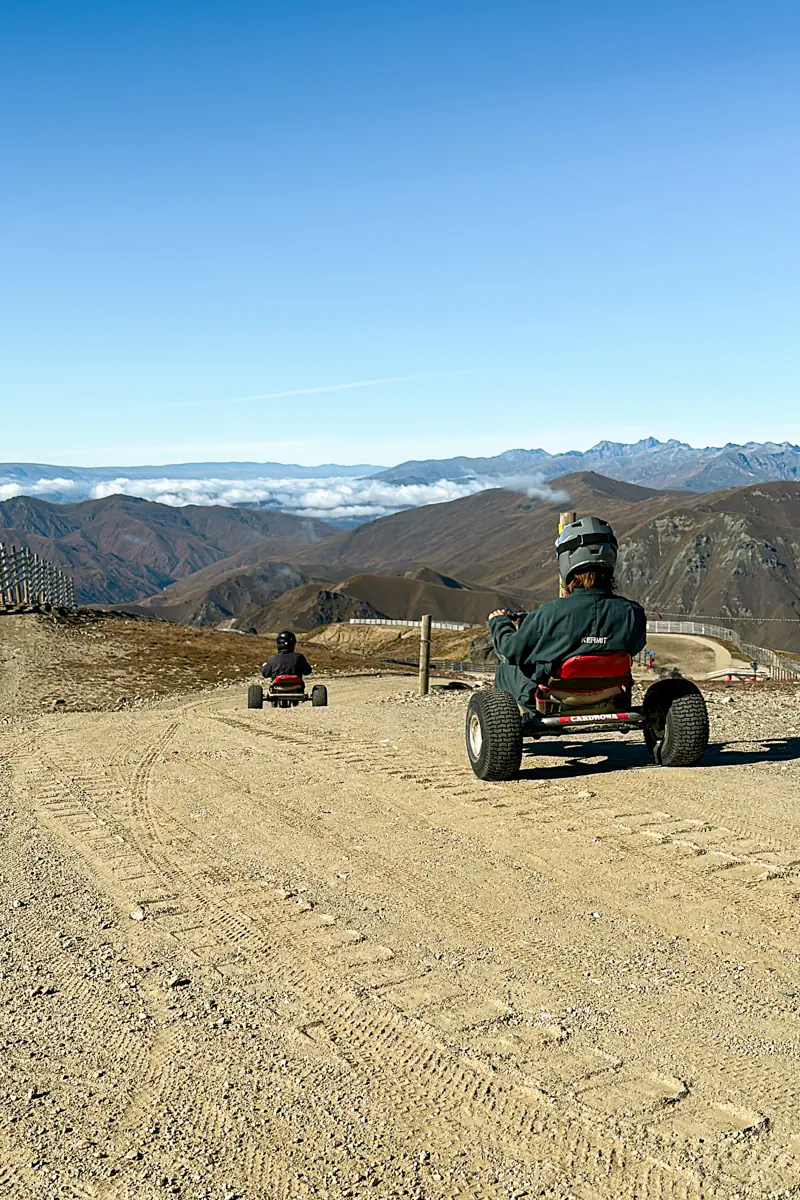 Two riders drive mountain carts downhill on a wide gravel track with sweeping alpine views in the background. The scene highlights the open landscape and sense of adventure at a high-altitude mountain resort in New Zealand.