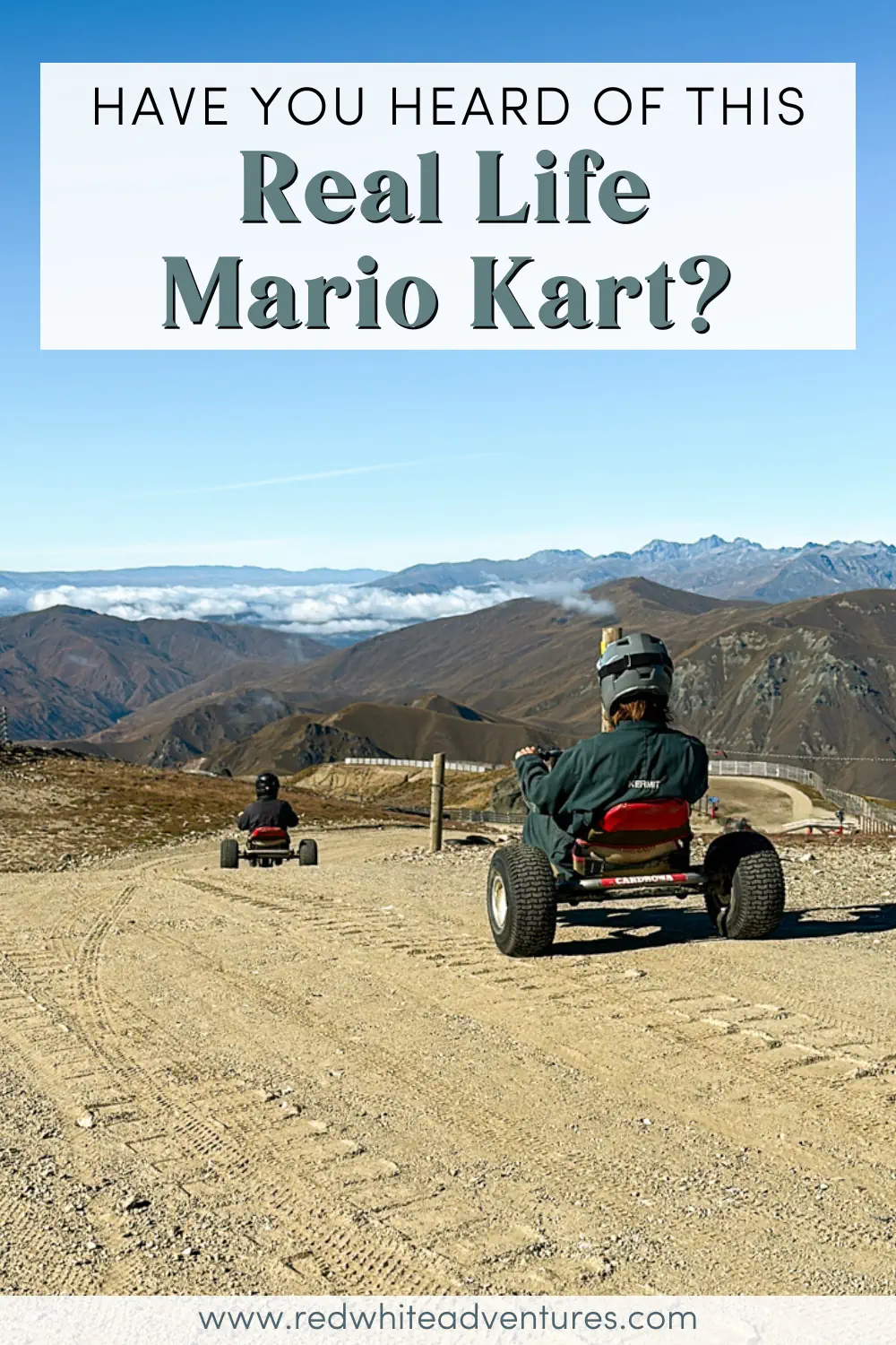 Two riders descend a wide gravel mountain cart track with sweeping alpine views stretching into the distance. The scene captures the thrill of downhill riding surrounded by rugged mountains and a clear blue sky.
