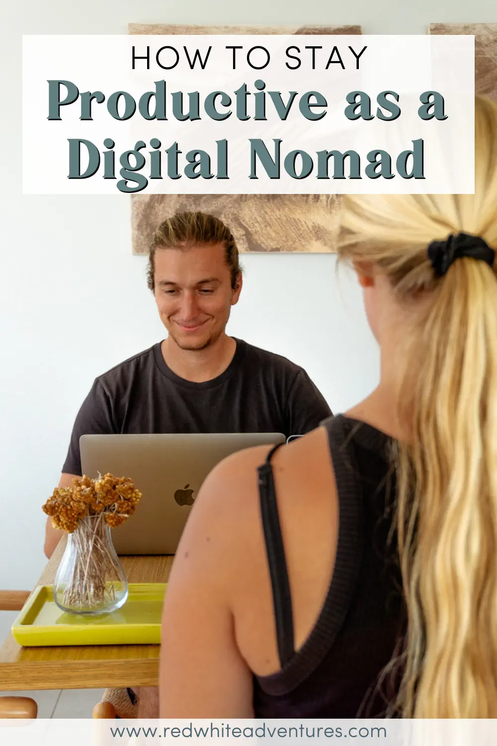 A man works on his laptop at a table while a woman sits across from him in a bright indoor workspace. The scene illustrates a calm and focused digital nomad setup.