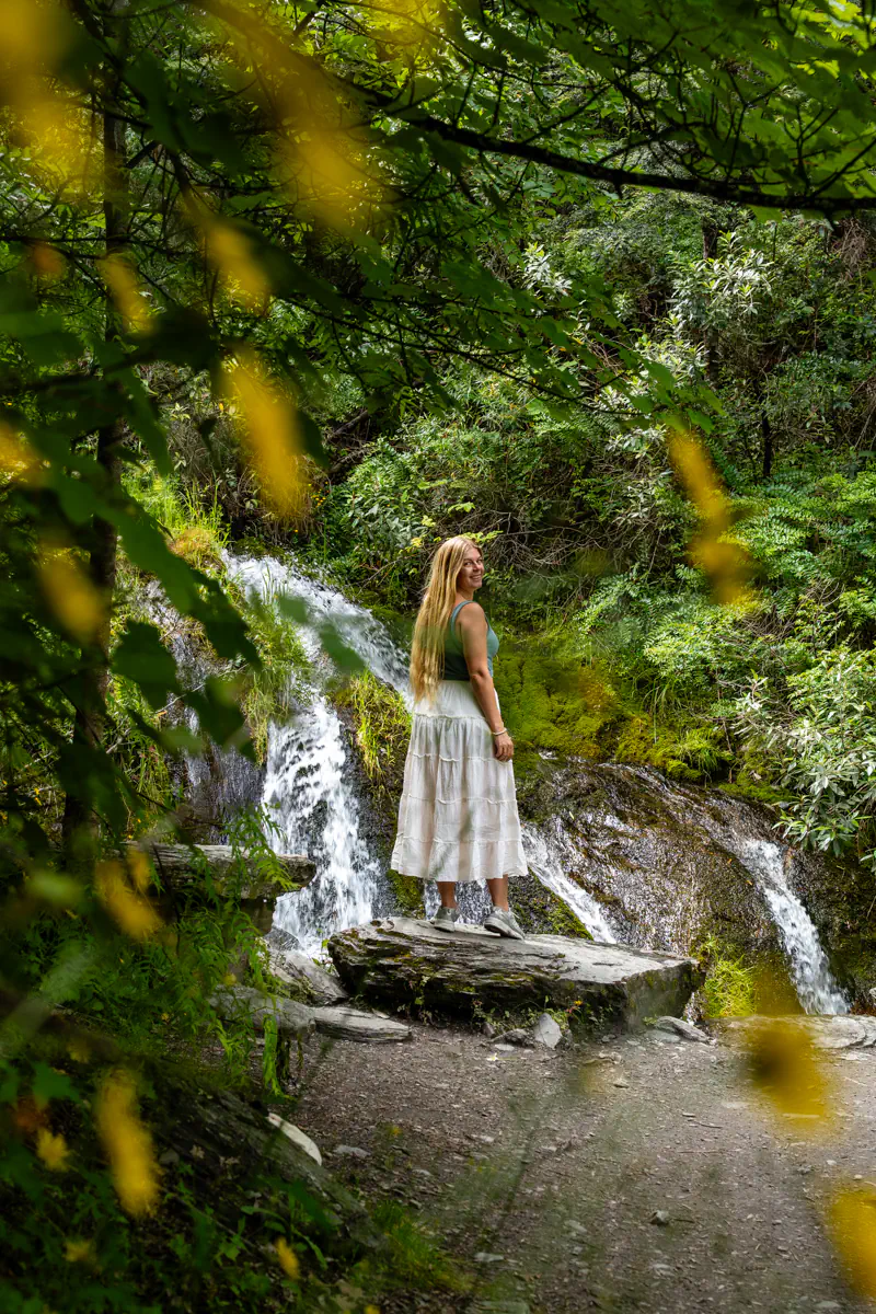 A woman stands on a rock beside a small waterfall, surrounded by lush forest and hanging branches. Soft light filters through the leaves, adding to the tranquil feel of the scene. The photo is framed by yellow, blurry flowers.