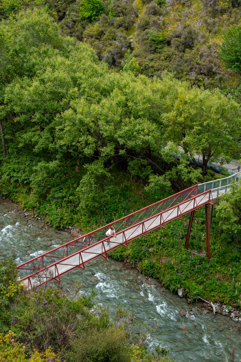 A red pedestrian bridge crosses a fast-moving river, surrounded by thick greenery and shrubs. A person pauses on the bridge, taking in the forested surroundings below.