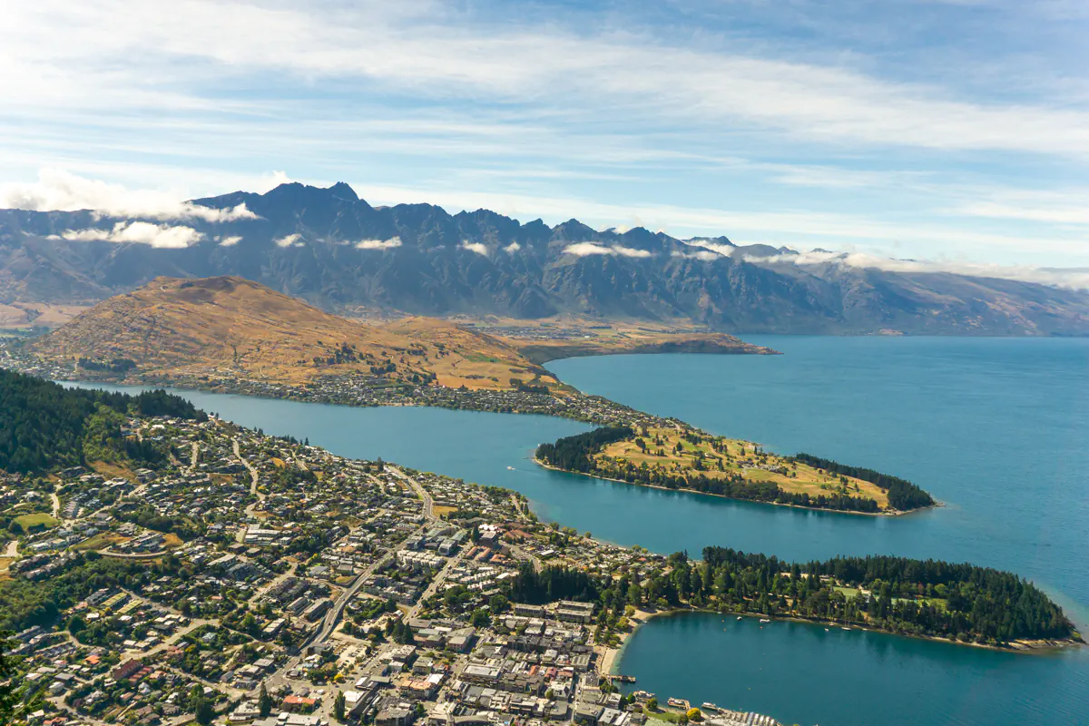 A panoramic view overlooks Queenstown and Lake Wakatipu from high above the town. The layered mountains and deep blue water highlight the scale of the Southern Alps landscape.