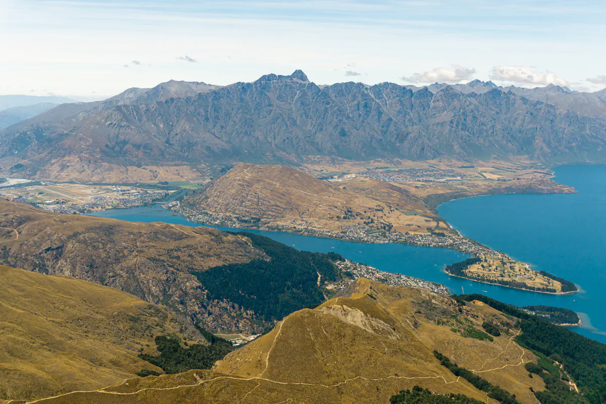 A wide aerial view looks down over Queenstown and Lake Wakatipu, with peninsulas, bays, and surrounding mountain ranges stretching into the distance. The blue water contrasts with golden hills and the town below.