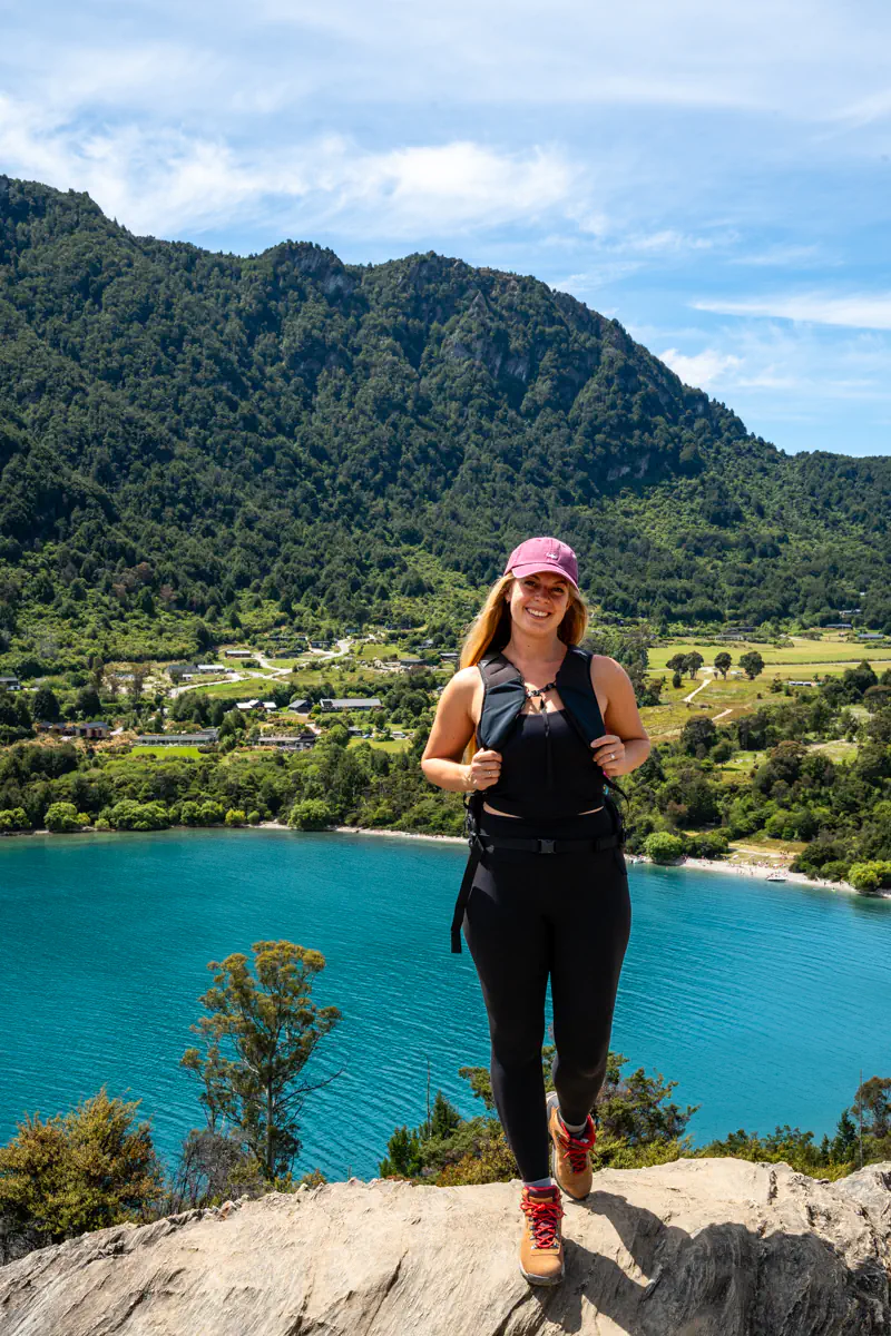 A woman stands on a rocky ledge overlooking a bright blue lake with a small beach and forested hills behind her. The clear water and sunny conditions emphasize the vibrant outdoor setting.