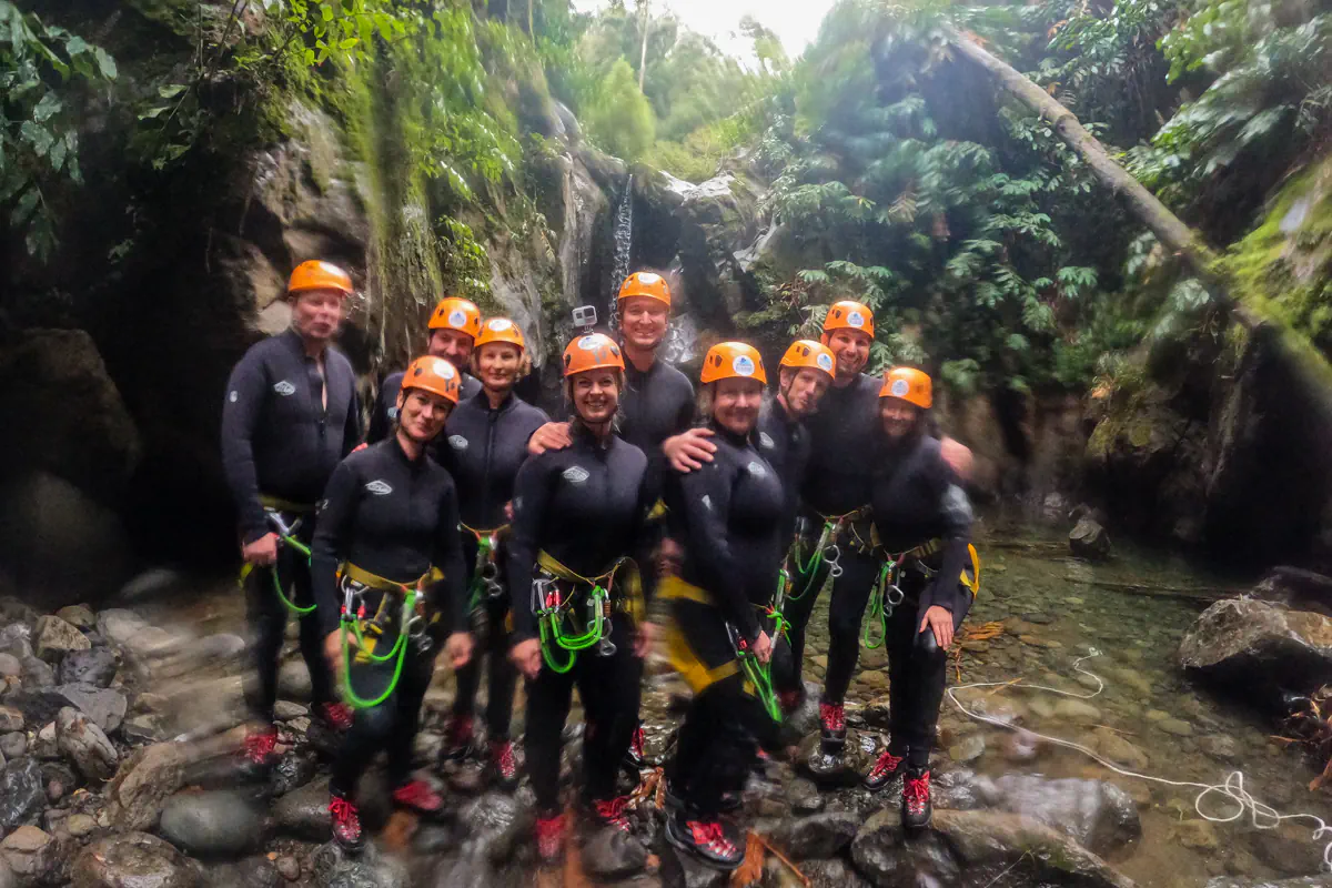 A group of canyoning participants pose together in wetsuits and helmets beside a small waterfall. The rocky canyon and flowing water create a dramatic natural backdrop.