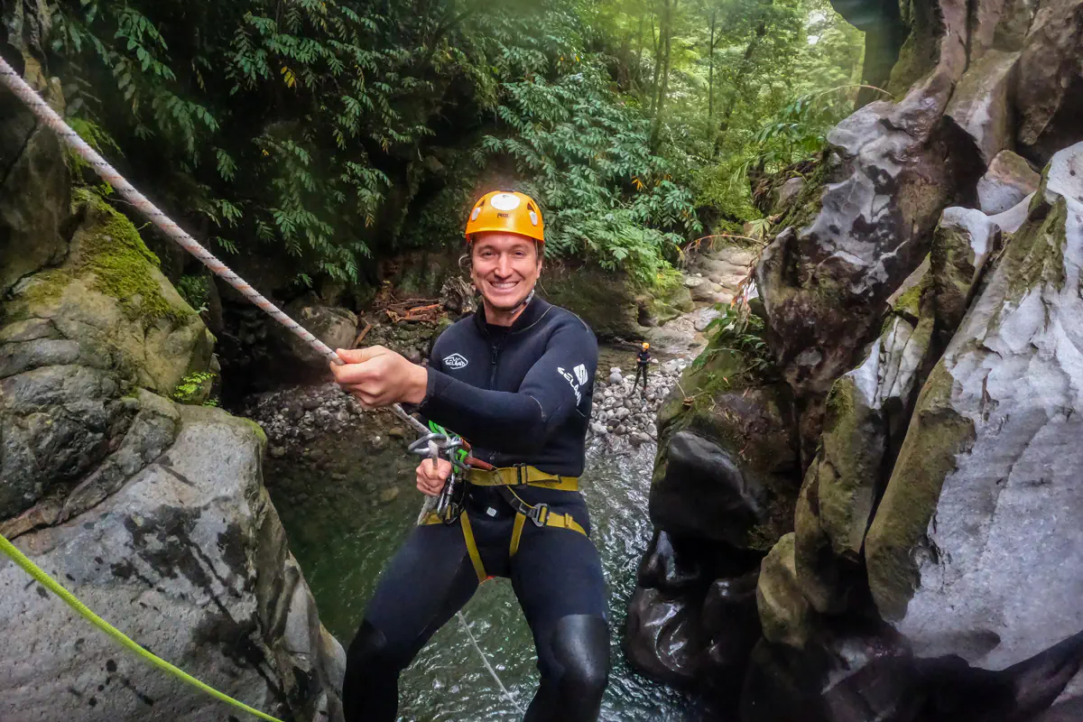 A man in a wetsuit and helmet rappels down a narrow canyon using a safety rope. A rocky stream and green vegetation are visible below him.