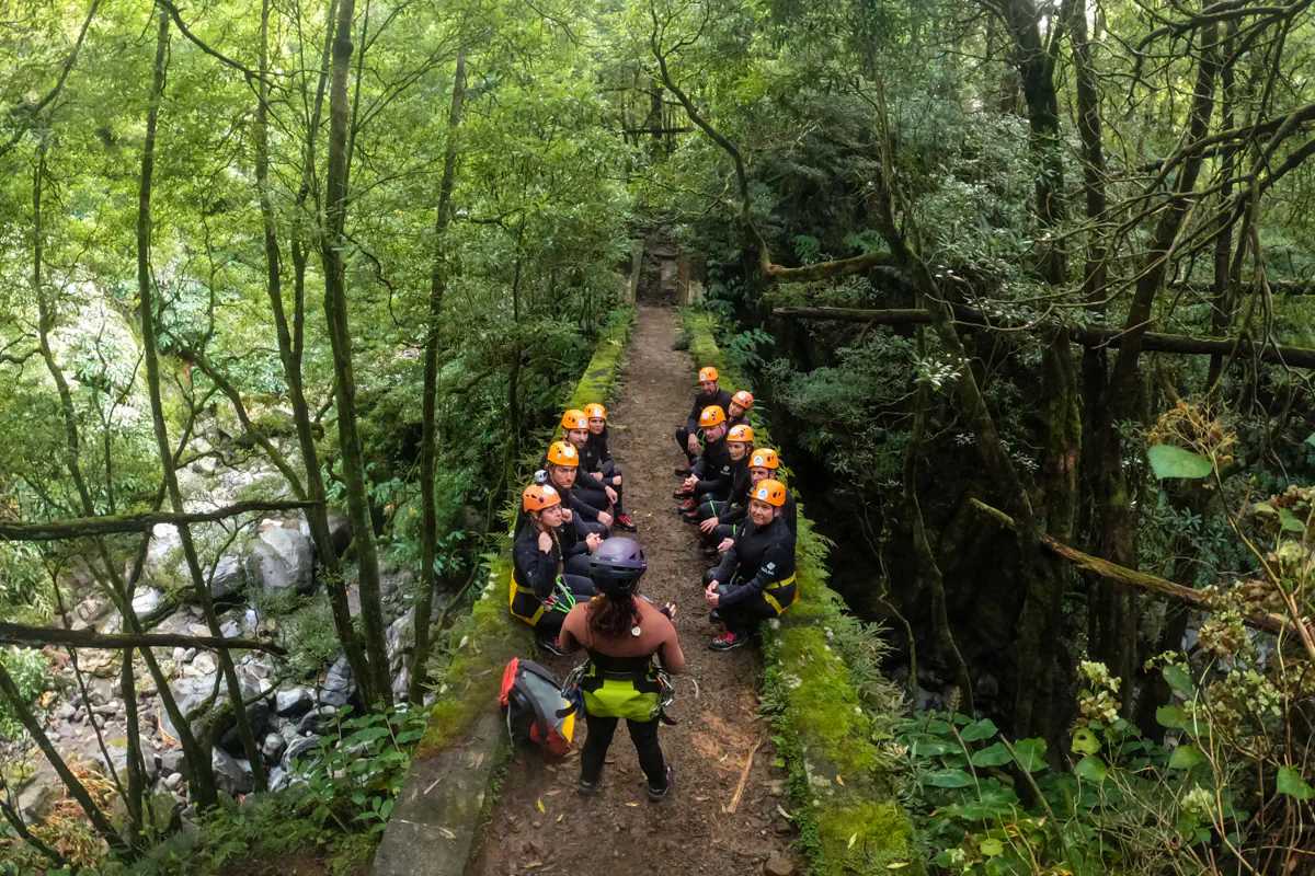 A group of canyoning participants sit on a mossy stone path in the forest during a briefing. They wear wetsuits, helmets, and harnesses surrounded by dense greenery.
