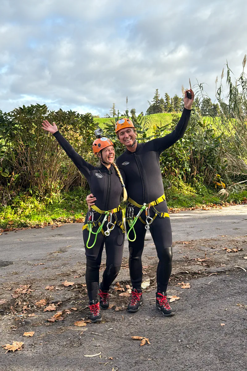A couple in canyoning gear celebrate with raised arms after finishing their adventure. Green fields and trees line the path behind them under a cloudy sky.