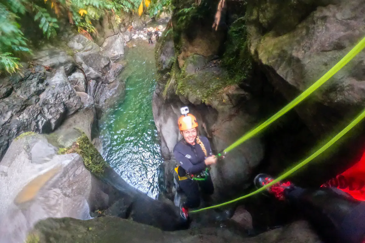 A woman in a helmet and wetsuit smiles while rappelling down a canyon wall beside a pool of water. Safety ropes stretch downward into the narrow gorge below.