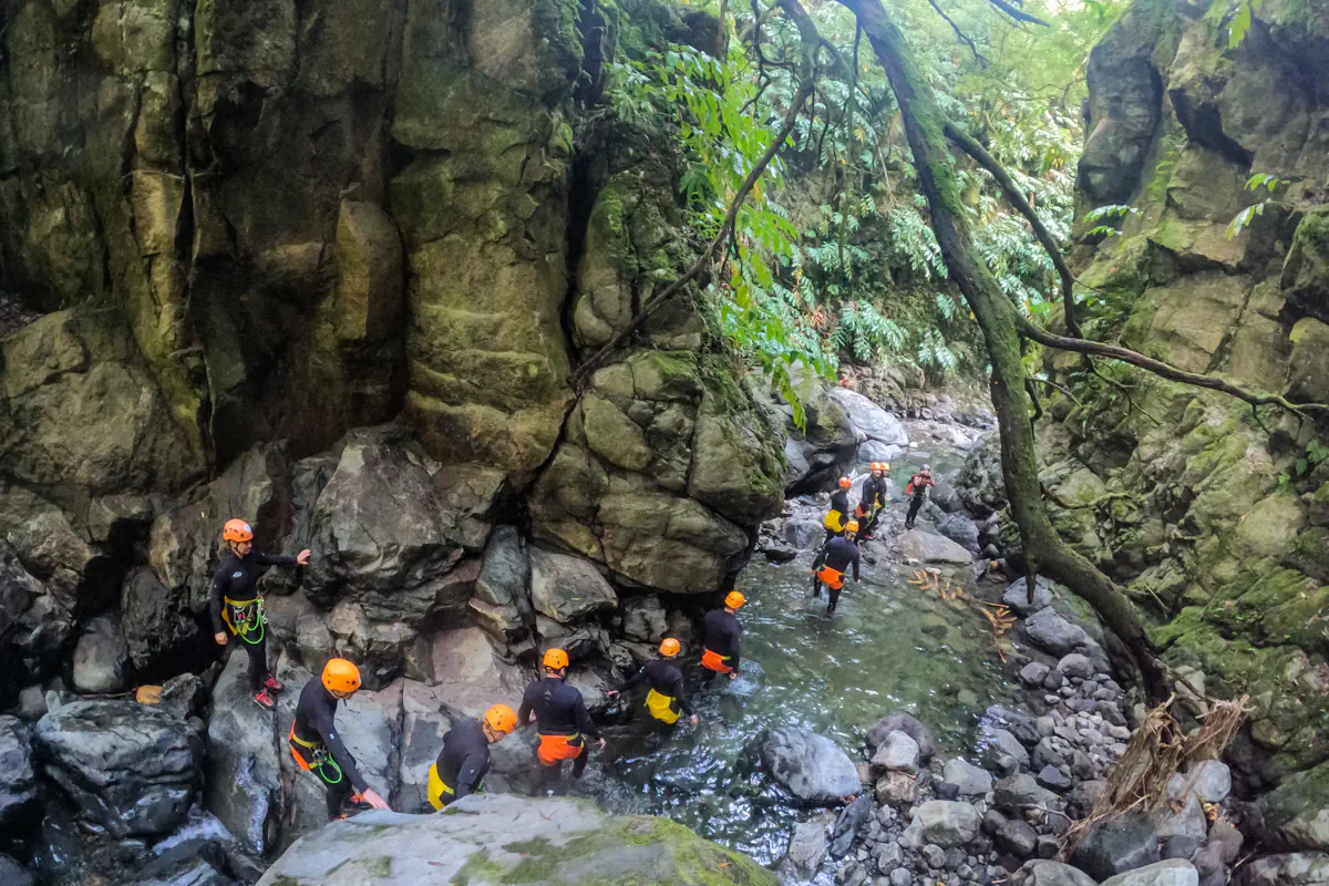 A group of canyoning participants wades through a rocky stream between steep canyon walls. Clear water flows through the gorge as the group carefully navigates the terrain.