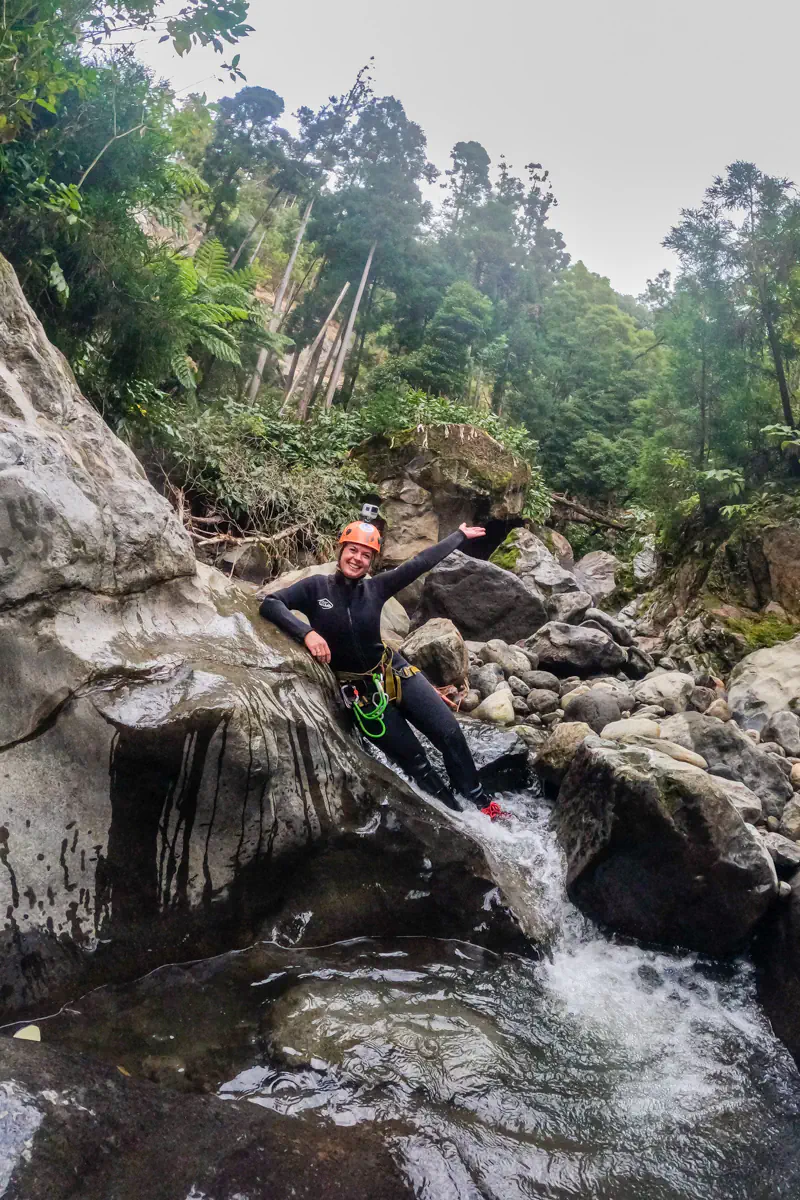A woman in canyoning gear smiles and gestures while laying on wet rocks beside flowing water. The lush forest and steep canyon walls create a dramatic natural setting.