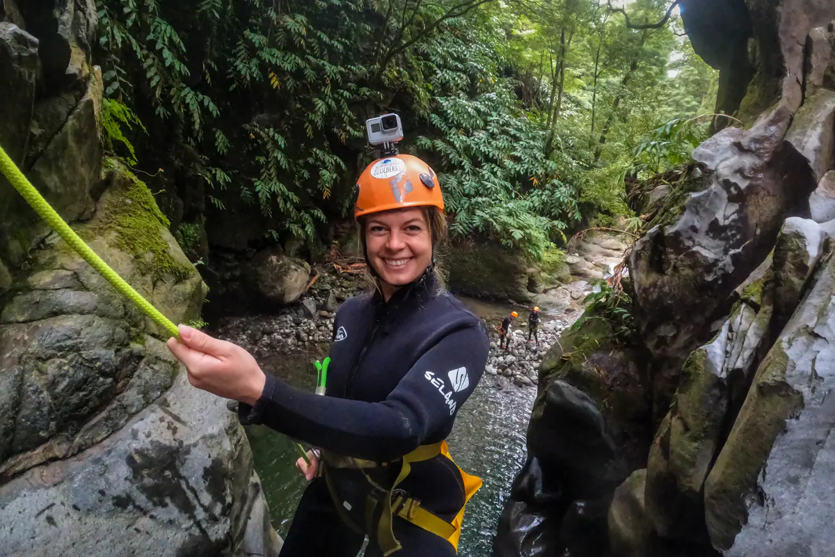 A woman wearing a helmet with an action camera rappels through a narrow canyon gap. Other canyoning participants wait below in the rocky stream.