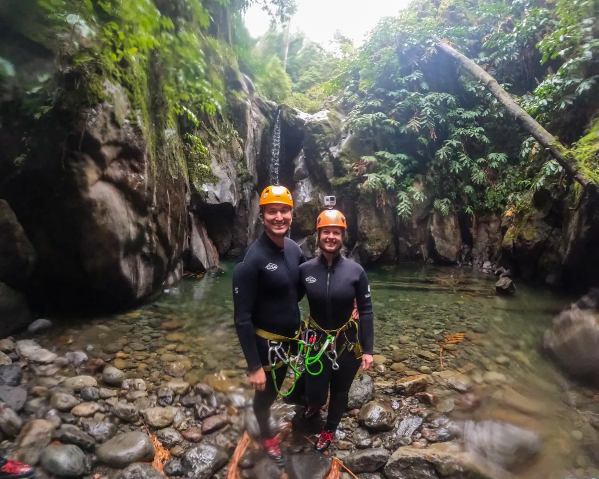 Two canyoning participants stand together in a shallow pool wearing helmets and harnesses. A small waterfall cascades behind them inside a narrow, green canyon.