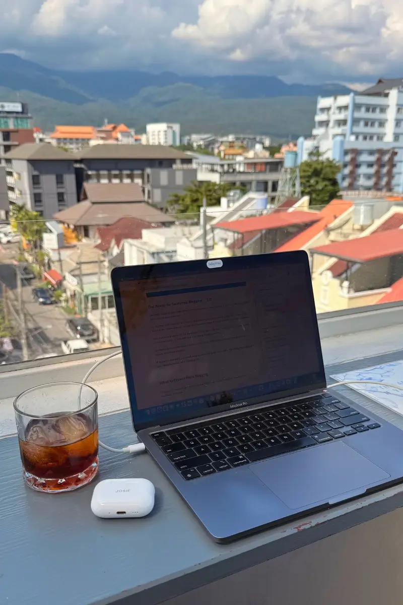 A laptop sits on a windowsill beside a glass of iced coffee and wireless earbuds, overlooking Chiang Mai city with mountains in the background. The image represents a digital nomad working remotely with an urban and mountain view.