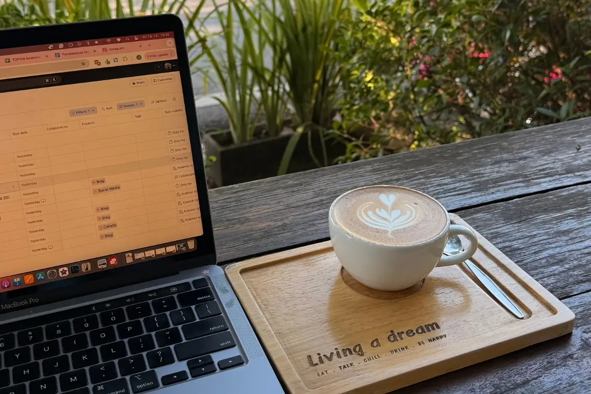 An open laptop sits on a wooden table next to a flat white coffee with heart-shaped latte art on a tray that reads “living a dream.” The photo captures a relaxed café work setup often associated with the digital nomad lifestyle.