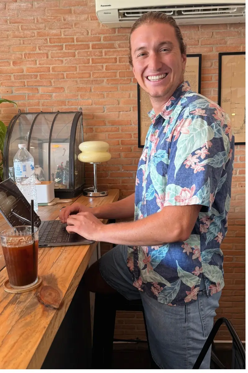 A smiling man works on a laptop at a wooden counter inside a café, with a brick wall and framed artwork behind him. The image captures a casual digital nomad workday.
