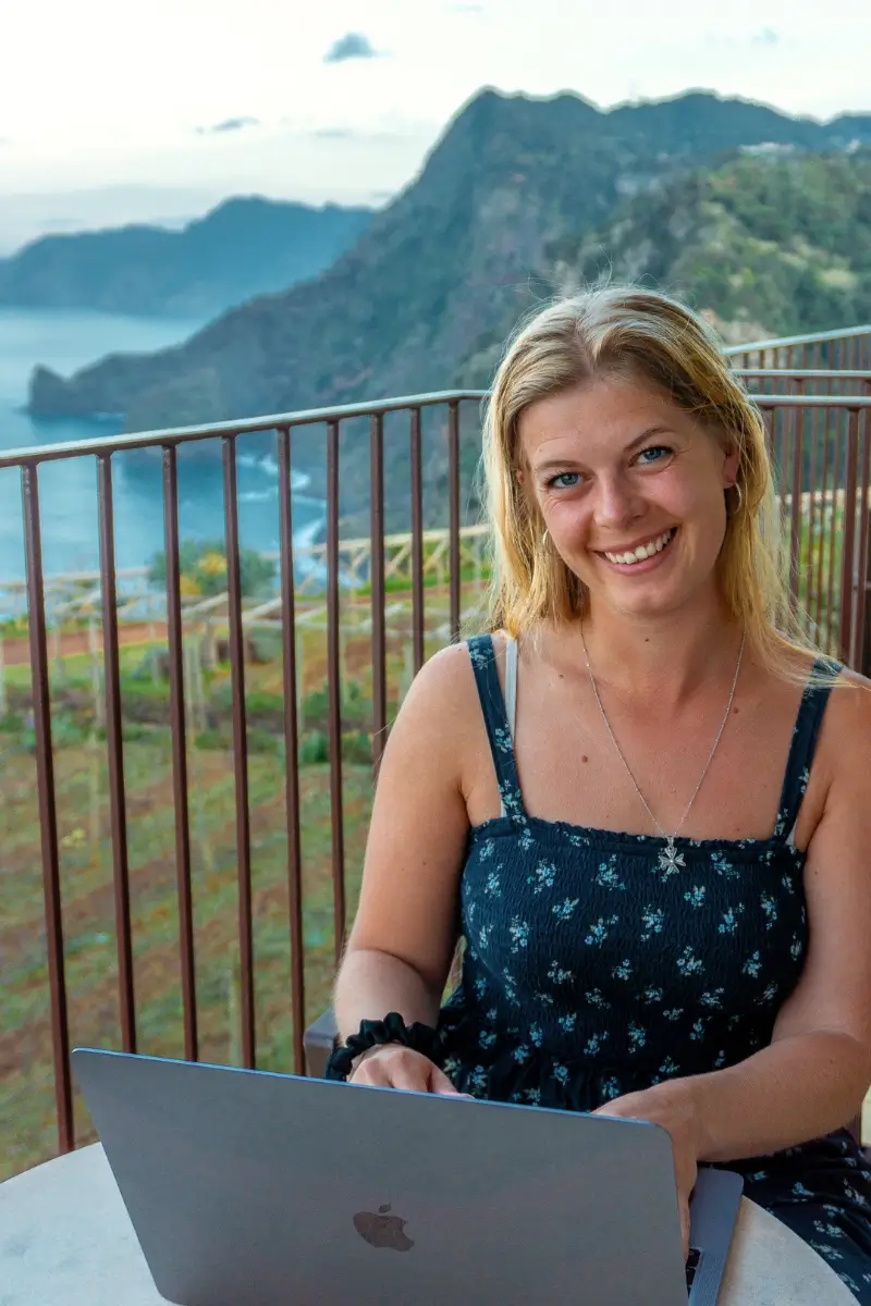 A woman smiles while working on a laptop at an outdoor table, with dramatic coastal cliffs and the ocean in the background. The image captures a relaxed digital nomad moment, blending remote work with a scenic seaside view.