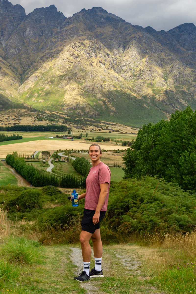 A man stands on a hiking trail holding a water bottle, smiling with dramatic mountain peaks rising behind him. The valley below shows farmland, winding roads, and patches of forest under shifting light.