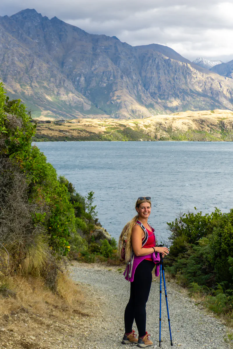 A woman pauses on a lakeside hiking trail with trekking poles, looking back toward the camera. Lake Wakatipu and steep mountain slopes form a striking backdrop beneath cloudy skies.