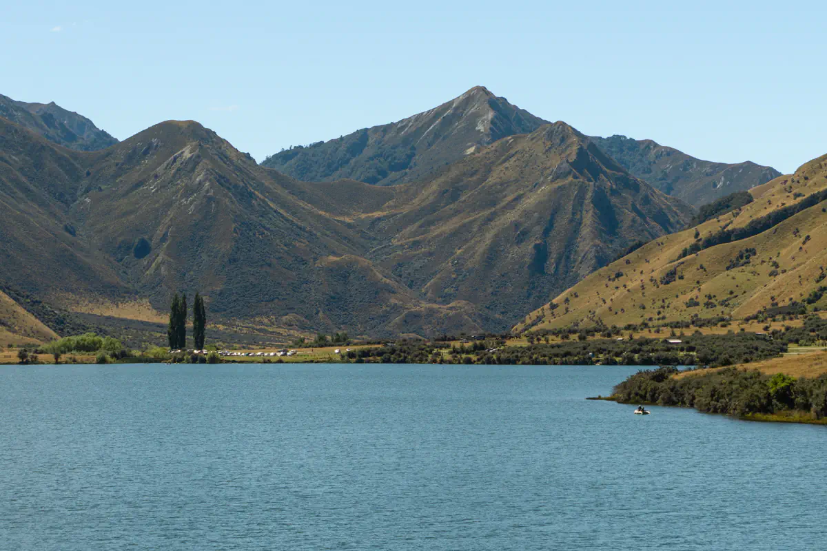 A calm blue lake sits beneath steep, rugged mountains covered in dry tussock grass in Central Otago, New Zealand. The still water reflects the quiet, remote feel of the alpine landscape.
