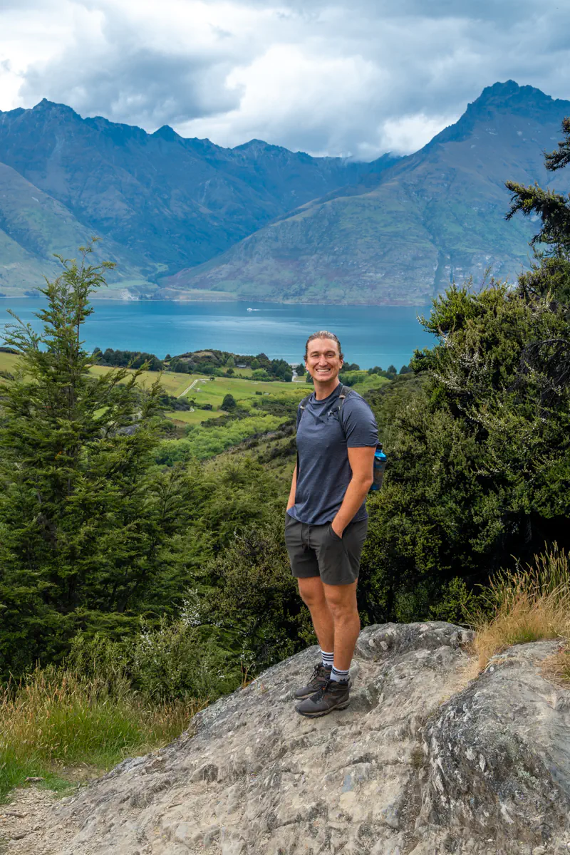 A hiker stands on a rocky lookout with Lake Wakatipu and dramatic mountain ranges stretching out behind him. Green hills and low clouds frame the expansive alpine landscape.