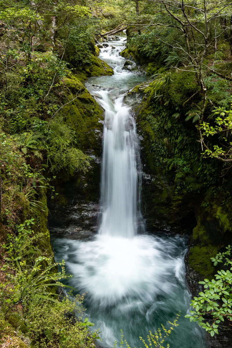 A narrow waterfall flows through a moss-covered gorge surrounded by dense green forest. The water cascades into a clear pool.