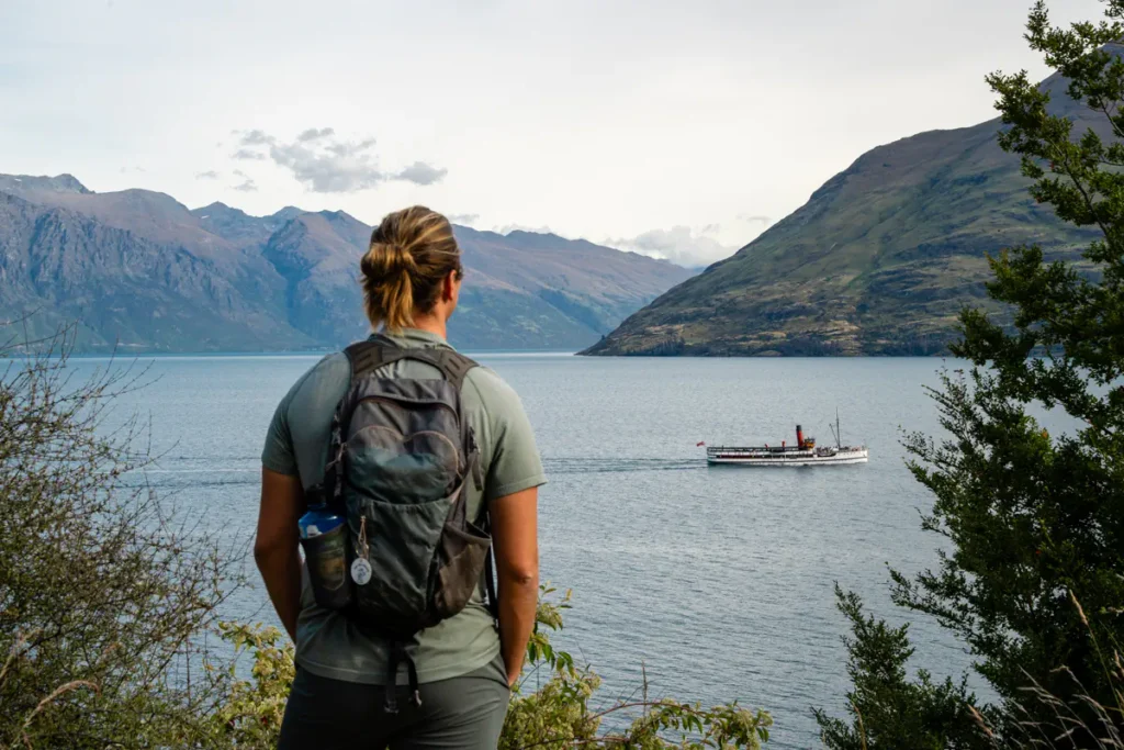 A hiker with a backpack looks out over Lake Wakatipu as a vintage-style boat cruises across the water. Mountains rise steeply from the shoreline, creating a calm yet dramatic view.