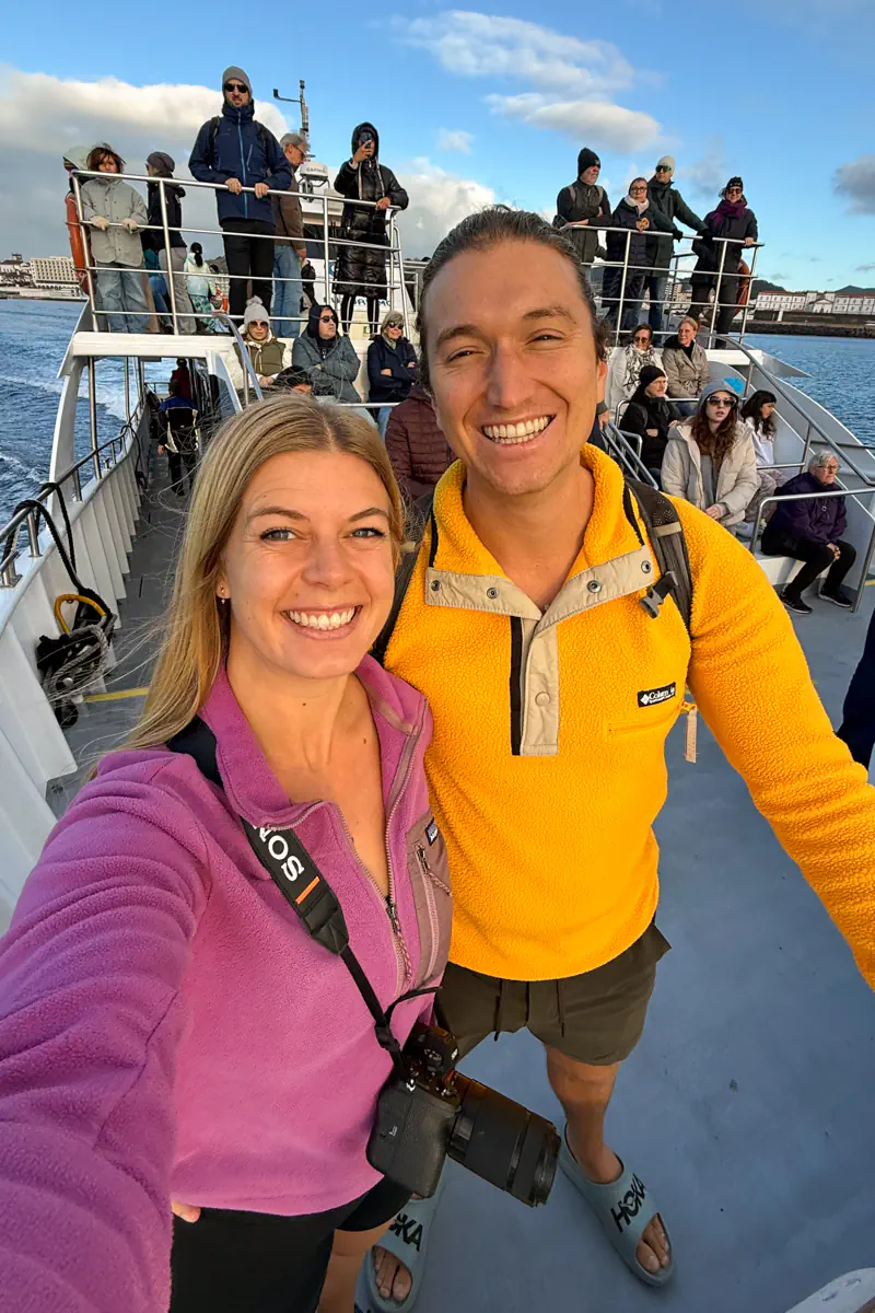 Two smiling travellers take a selfie on the deck of a catamaran in Ponta Delgada, with other passengers gathered behind them. The ocean and harbour are visible under a bright, partly cloudy sky.