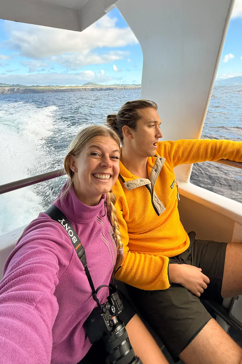 Two travellers sit on a whale-watching boat as it moves through the Atlantic Ocean near Ponta Delgada. Waves trail behind the boat, with the coastline of São Miguel Island visible in the distance. The traveller on the left is smiling while the traveller on the right is yawning looking rather seasick.