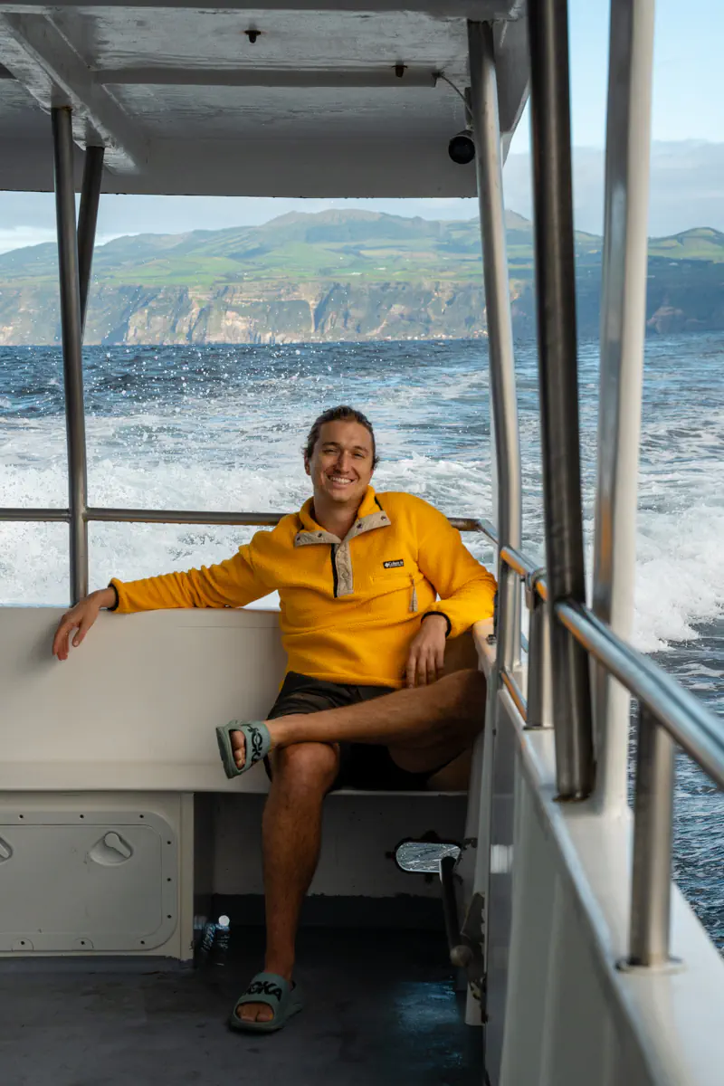 A smiling traveller sits relaxed on a small boat during a dolphin-watching trip on the Atlantic Ocean. Ocean spray and distant cliffs are visible behind them.