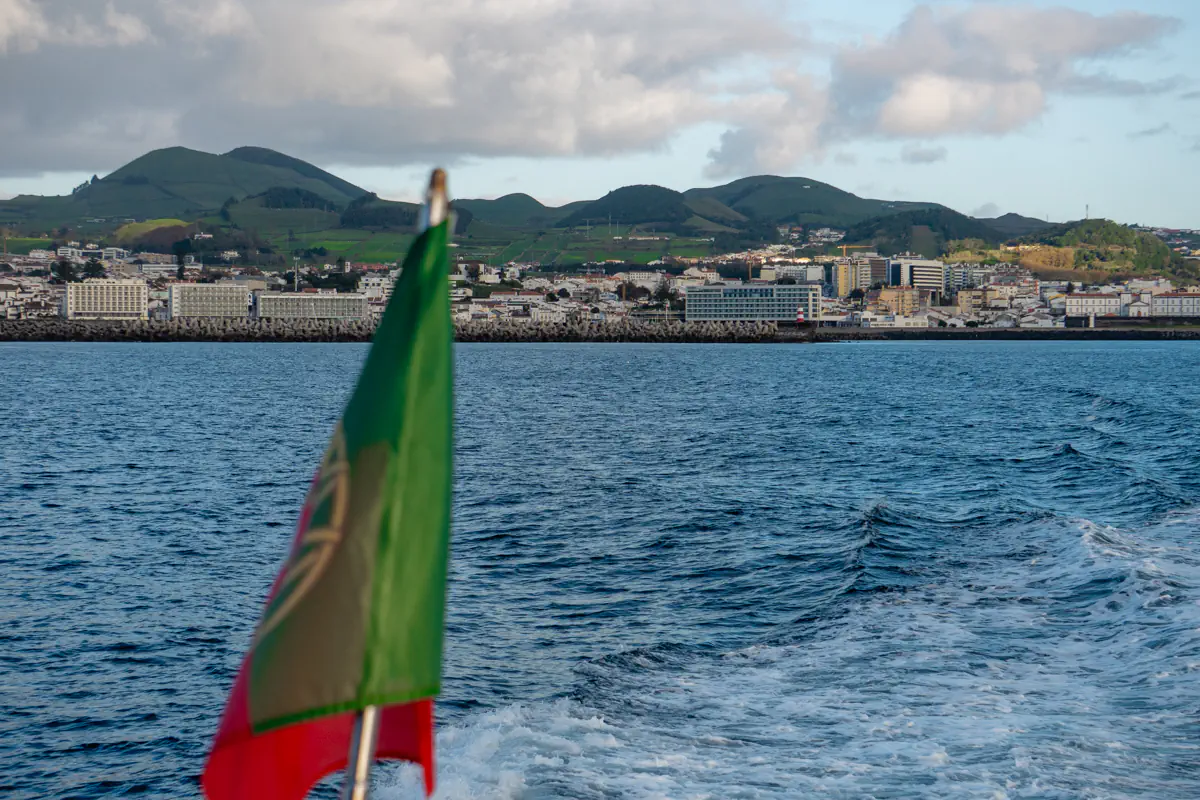 A boat heads away from the harbour, with a green and red Portuguese flag visible in the foreground. A coastal town and rolling green hills line the shoreline in the distance.