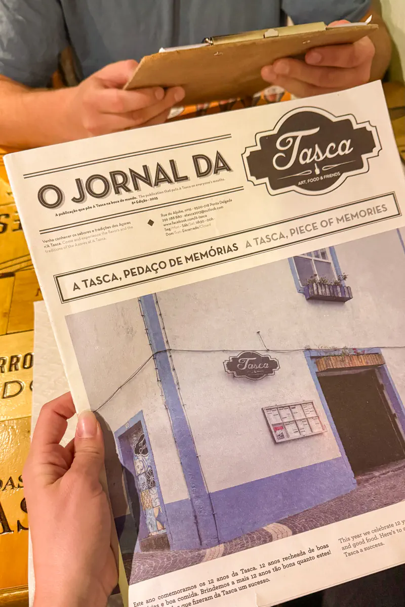 A person holds a newspaper titled “O Jornal da Tasca” featuring a photo of a building with a Tasca sign. A clipboard and another diner’s hands are visible across the table.