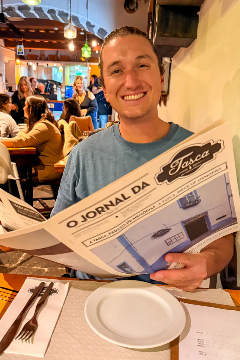 A smiling man sits at a restaurant table holding the “O Jornal da Tasca” newspaper menu. Other diners and hanging lights are visible inside the busy restaurant behind him.