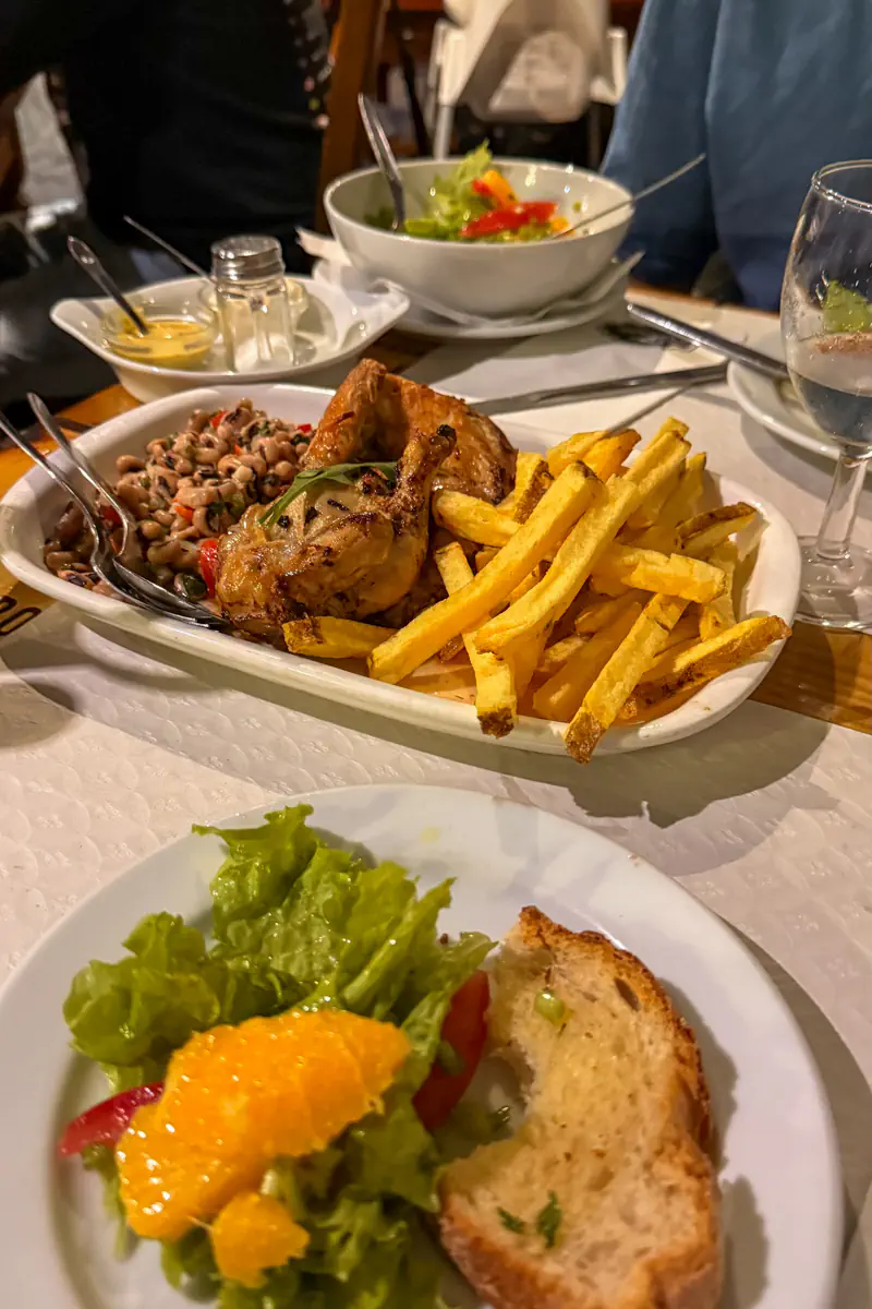 A table is set with a plate of grilled chicken, fries, and beans, along with salad, bread, and glasses of water. Multiple dishes and cutlery are visible, suggesting a shared meal.