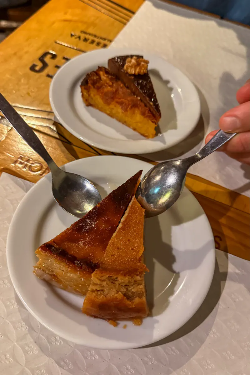 Two small plates hold slices of cake while hands with spoons reach toward one of the desserts. The plates rest on a wooden table with napkins and cutlery nearby.
