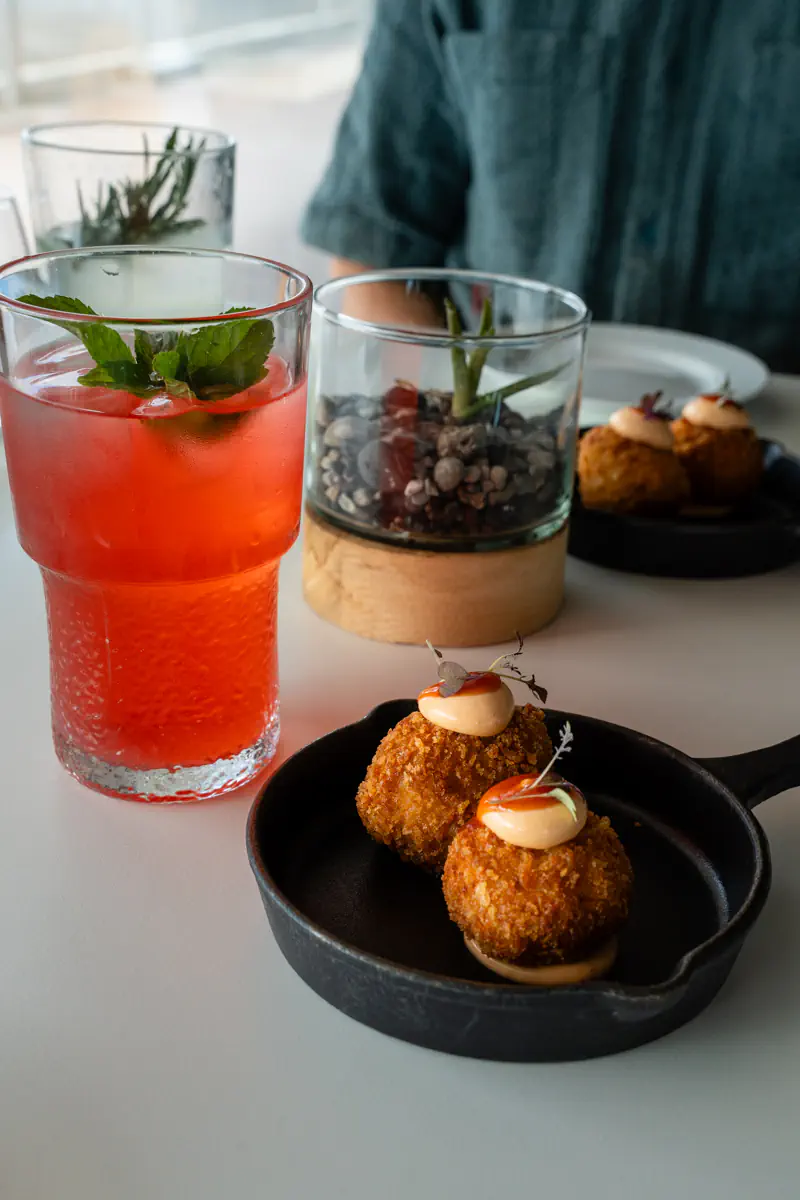 A pink lemonade garnished with fresh mint sits beside two golden-brown croquettes topped with a creamy mayonnaise in a small black pan. The dishes are placed on a light-coloured table with minimalist decor in the background.
